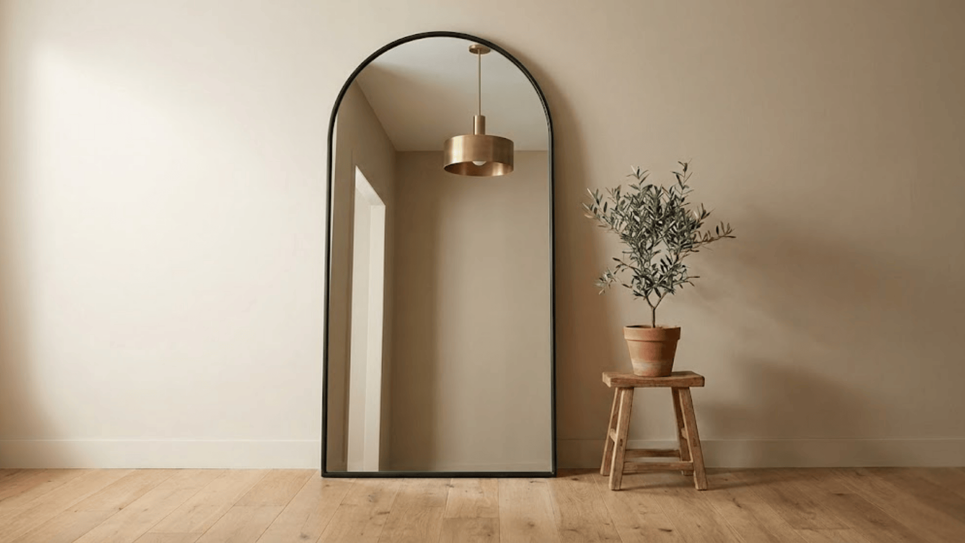 full length arched mirror in black metal frame leaning against beige wall with potted olive tree
