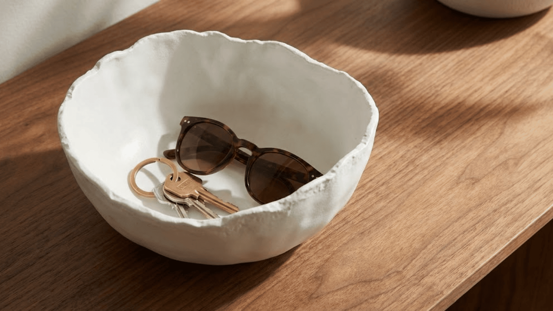 organic white ceramic bowl holding keys and sunglasses on walnut console surface with sculptural form