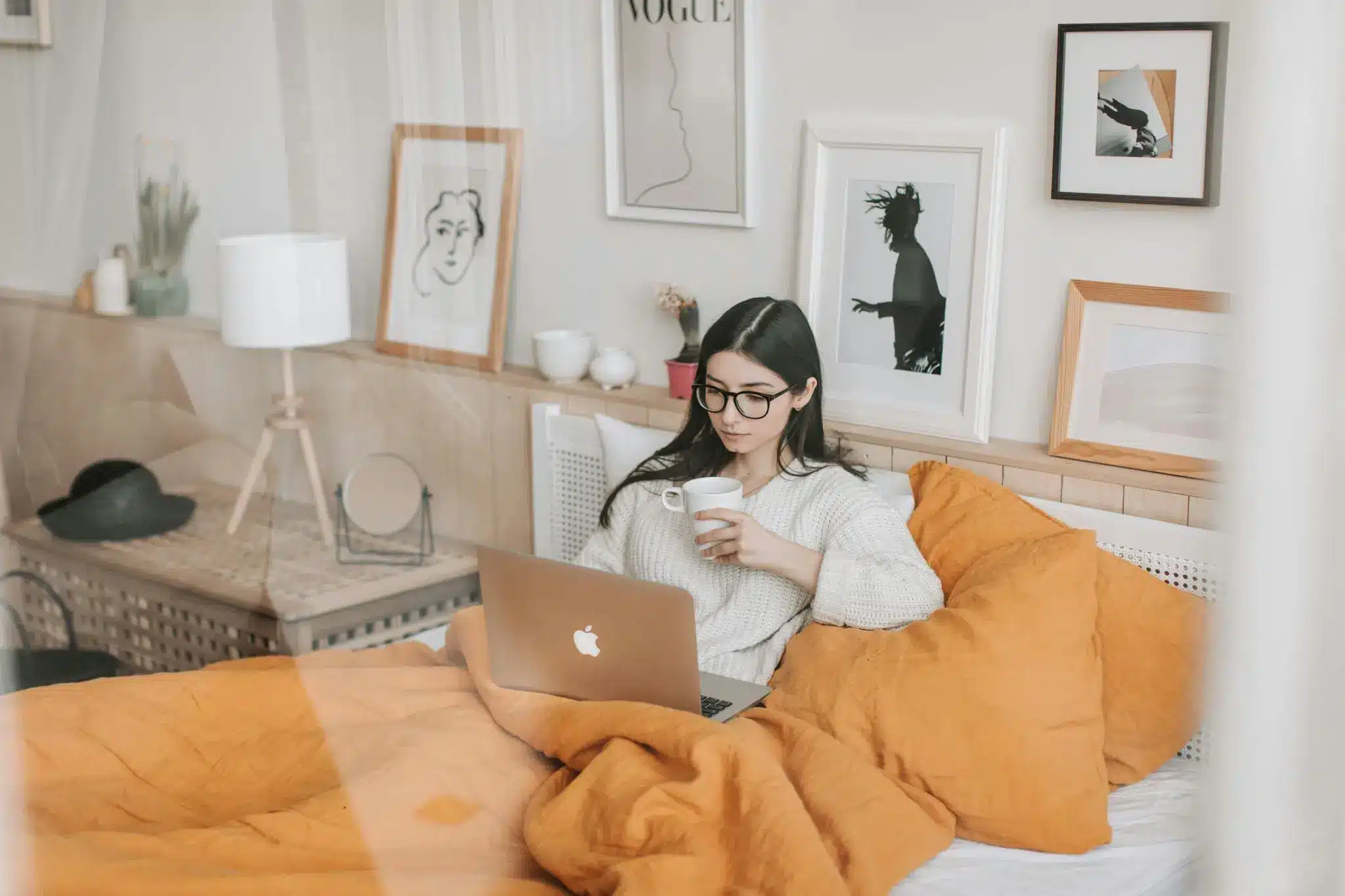 Woman in cozy bedroom working on laptop with coffee, surrounded by wall art and warm-colored bedding