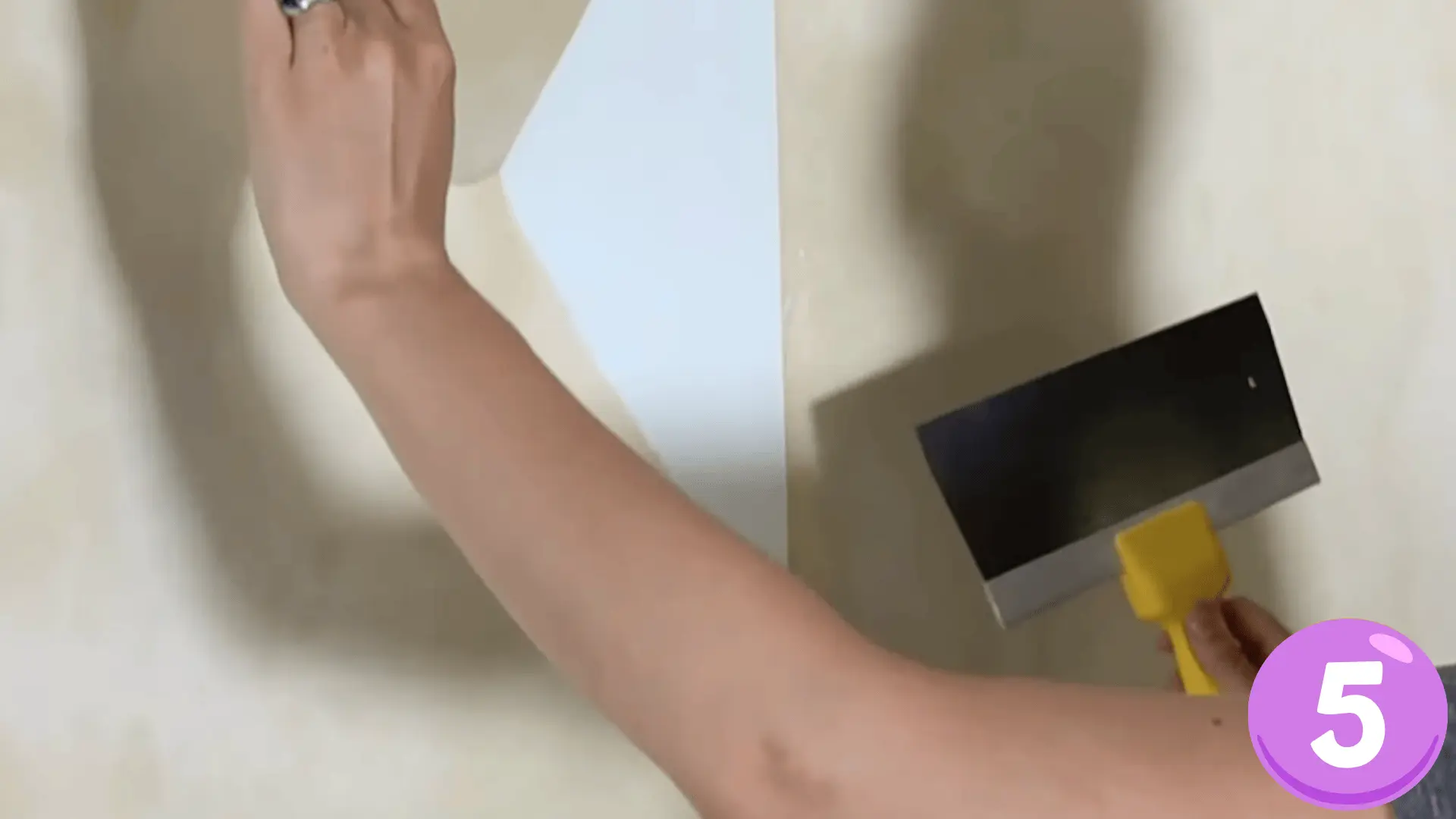 a person scraping off loosened wallpaper from a wall using a wide scraper with a yellow handle