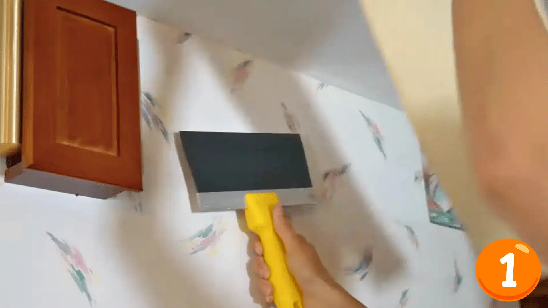a person using a wide scraper with a yellow handle to peel wallpaper off a wall