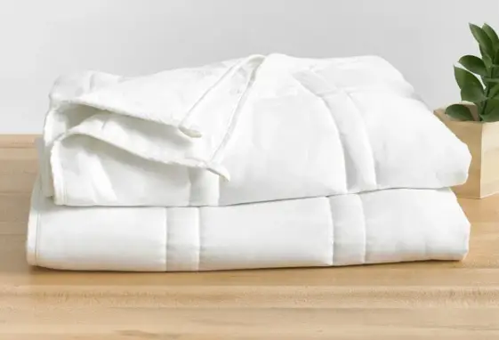 Folded white comforter on wooden table beside small potted plant in bright room