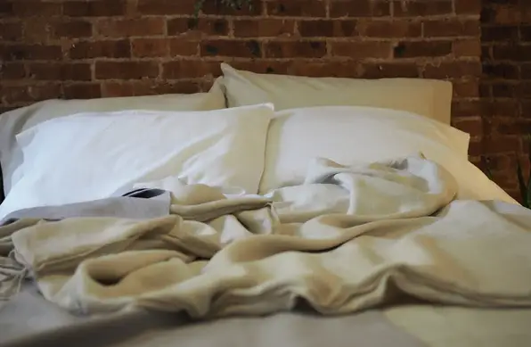 Untidy bed with white pillows and beige blankets against exposed brick wall