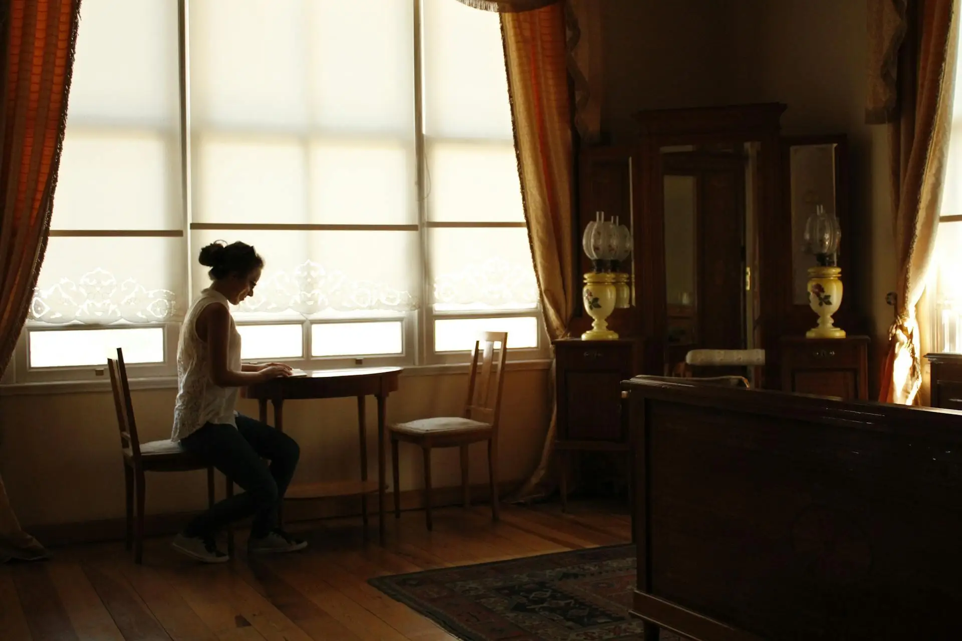Person seated at small table in dimly lit vintage room with lace curtains and antique furniture