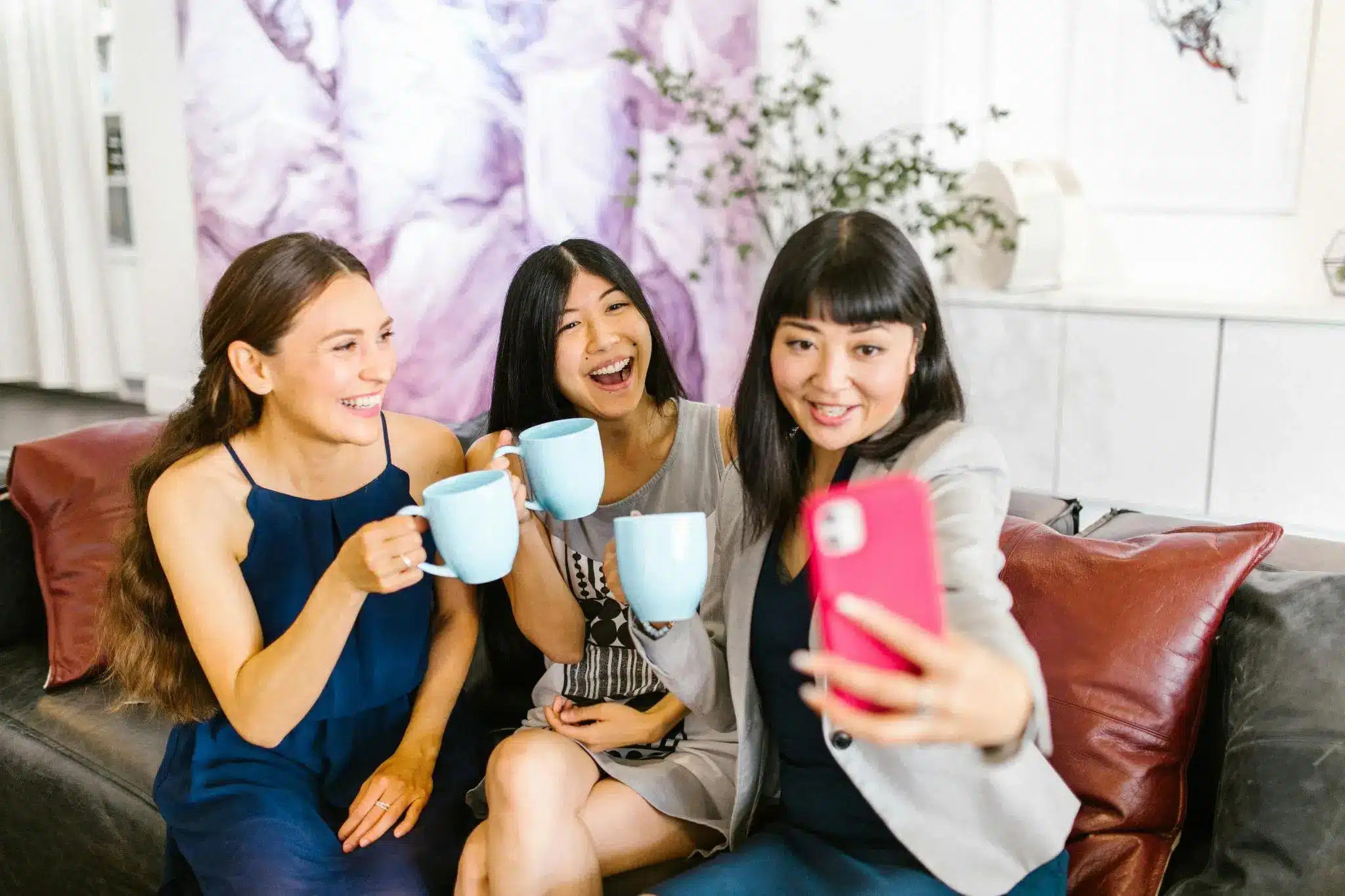 Three women sitting on a couch, holding mugs and taking a selfie indoors