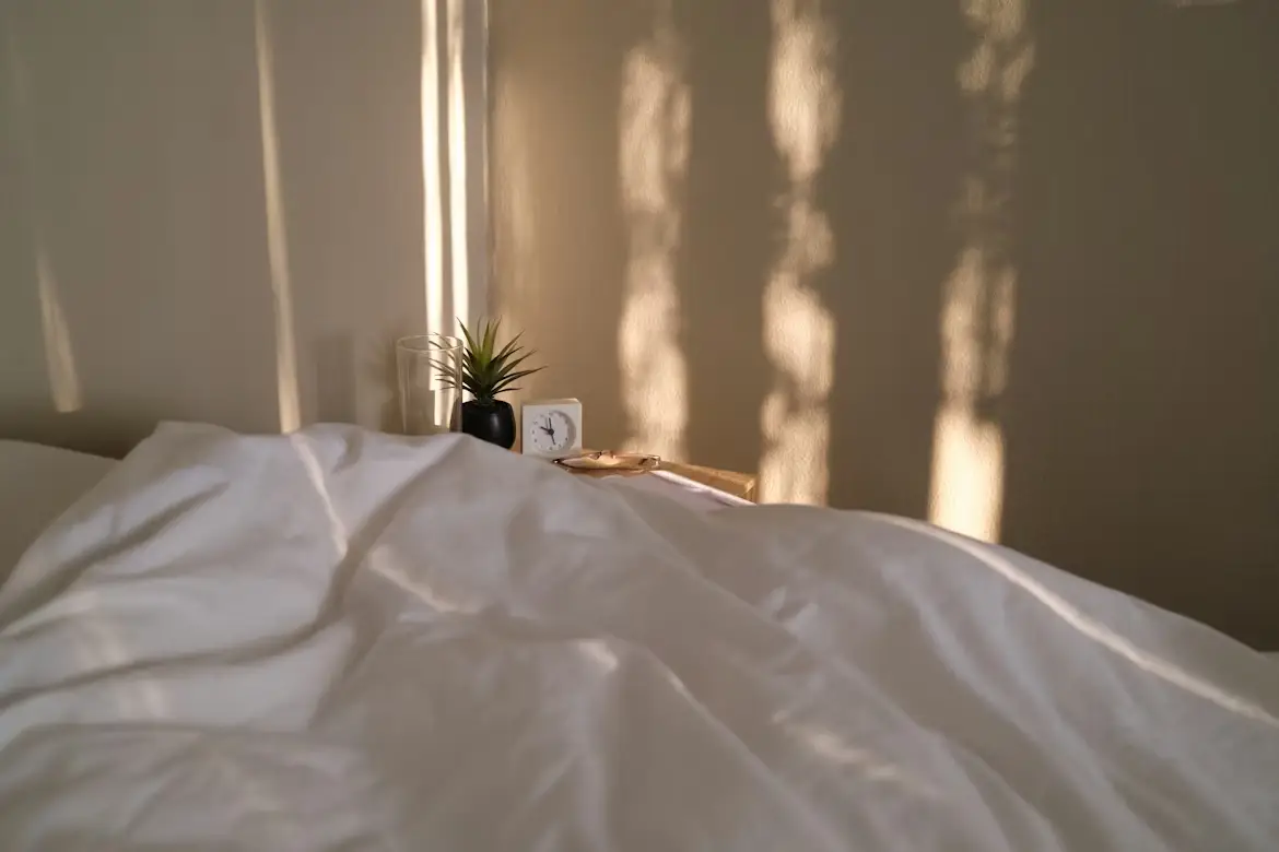 Bed with white sheets and morning light on wall, bedside table with clock and plant