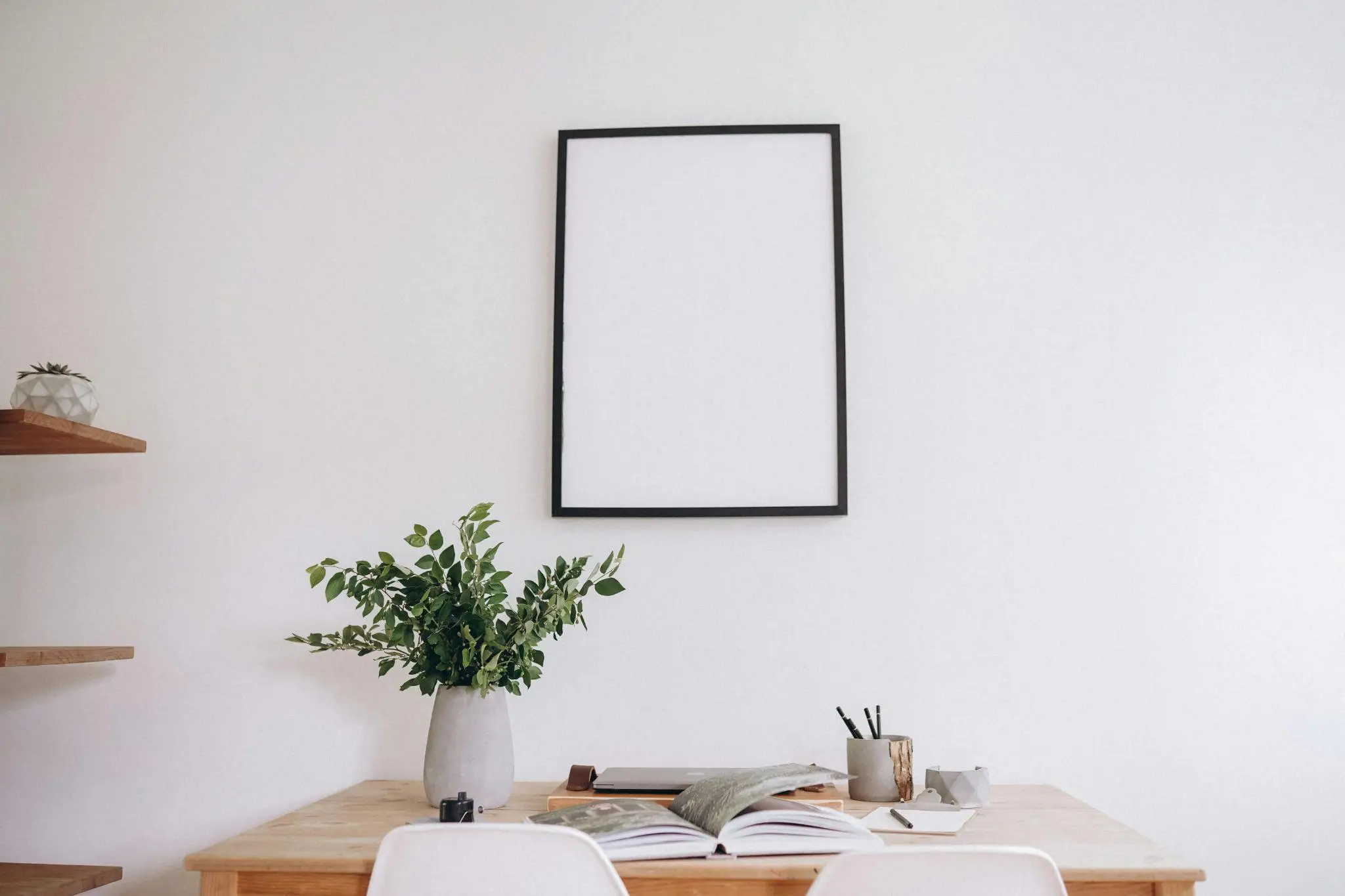 Empty black picture frame on white wall above wooden desk with plant and open book