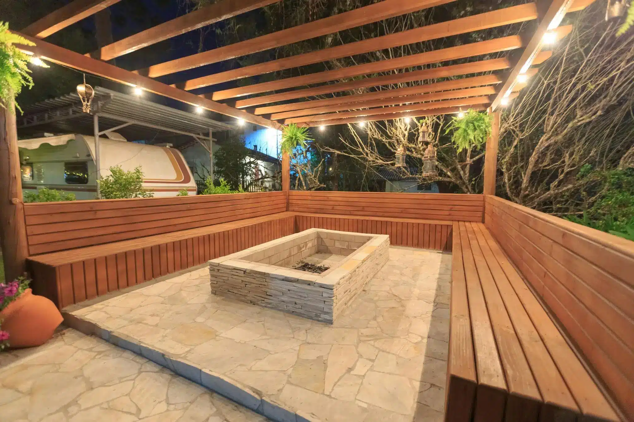 Outdoor wooden pergola seating area with central stone fire pit and ambient evening lighting