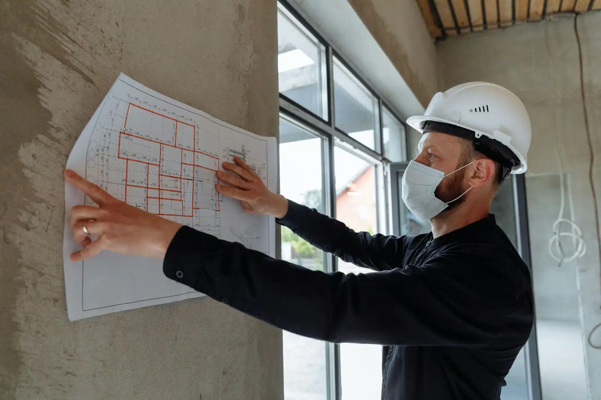 Architect wearing hard hat and mask examining blueprints on construction site wall