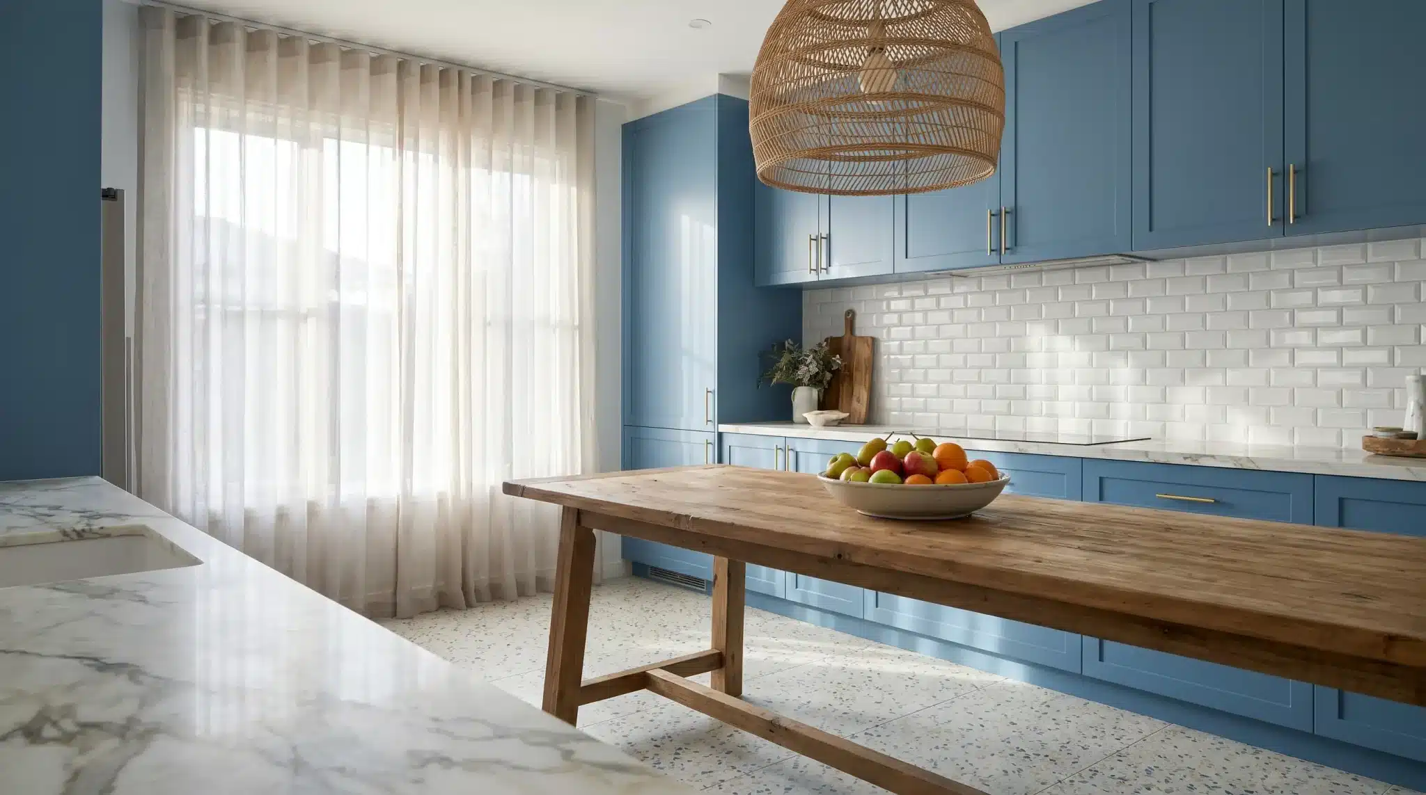 Wooden kitchen table with fruit bowl, blue cabinets, and woven light fixture in bright kitchen