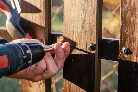 Person using power drill to fasten screws into wooden fence outdoors in sunlight