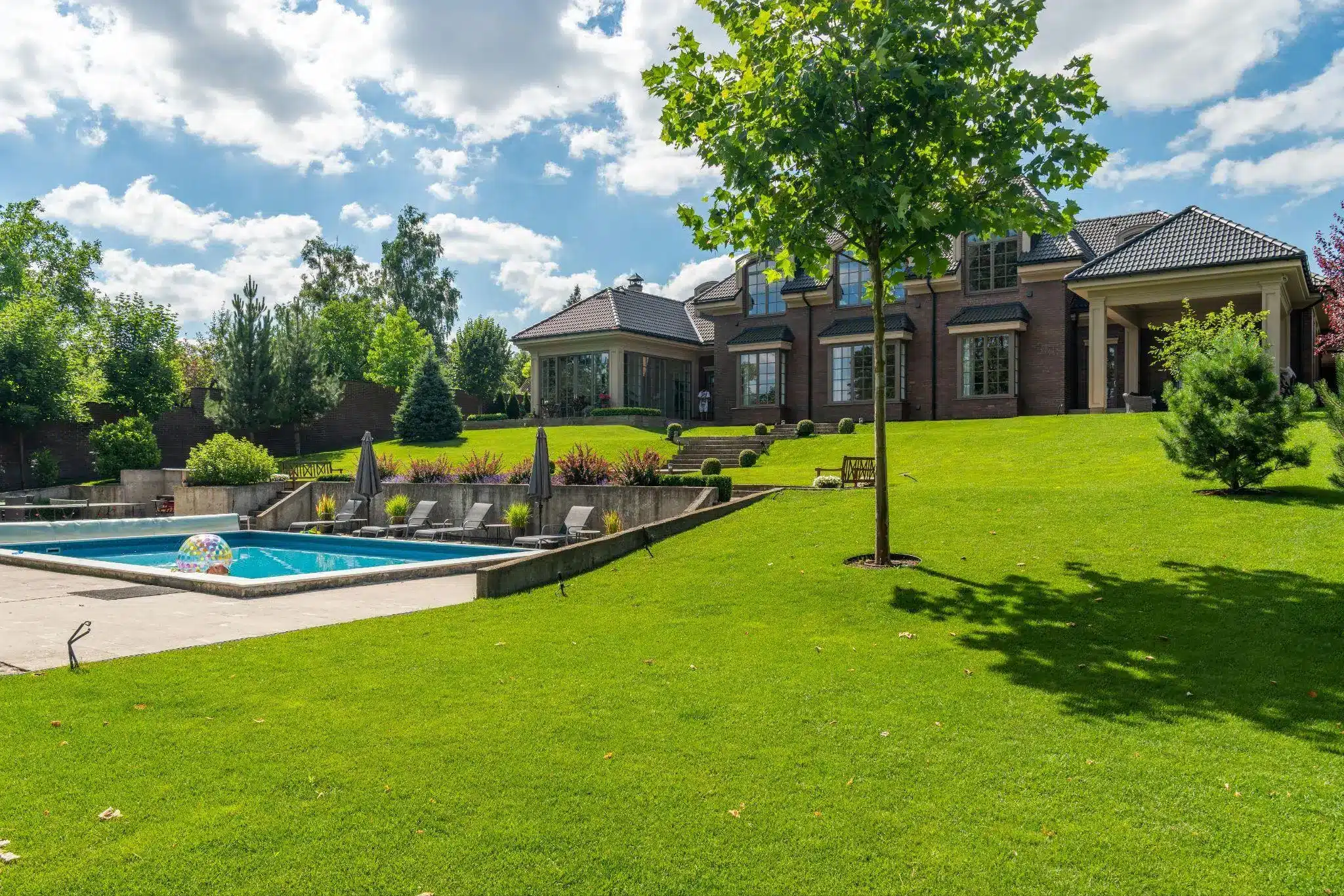Lush green lawn leading to a brick house with a swimming pool and sun loungers