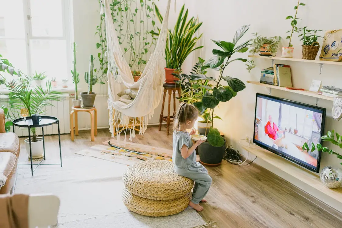 Child sitting on woven pouf eating and watching TV in plant-filled living room