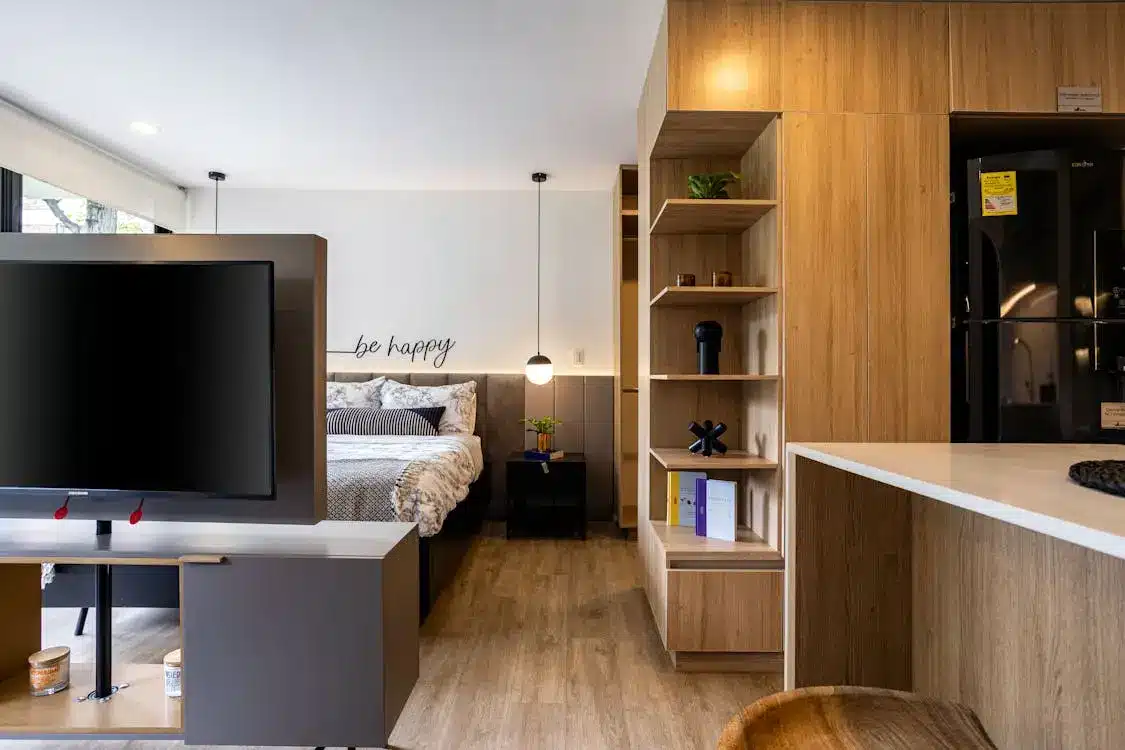 Modern studio apartment with bed, shelves, TV, and wooden cabinetry in bright setting