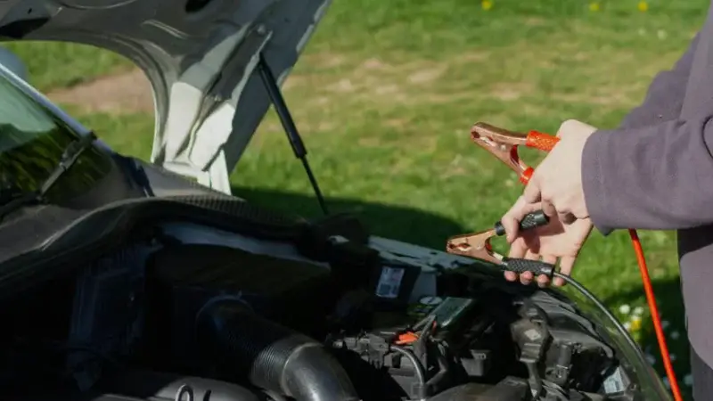 Person holding jumper cables near car engine in outdoor setting with green grass background