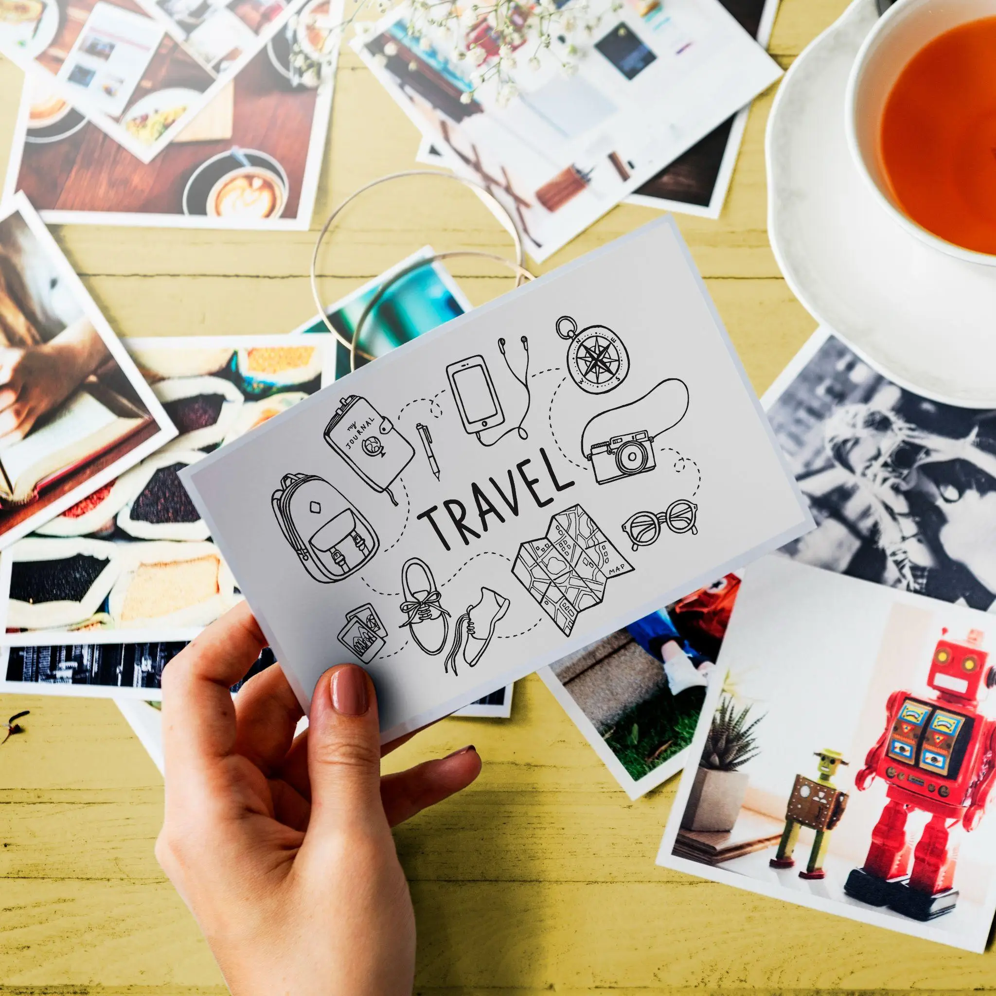 Hand holding travel-themed postcard with doodles, surrounded by photos and cup of tea on table