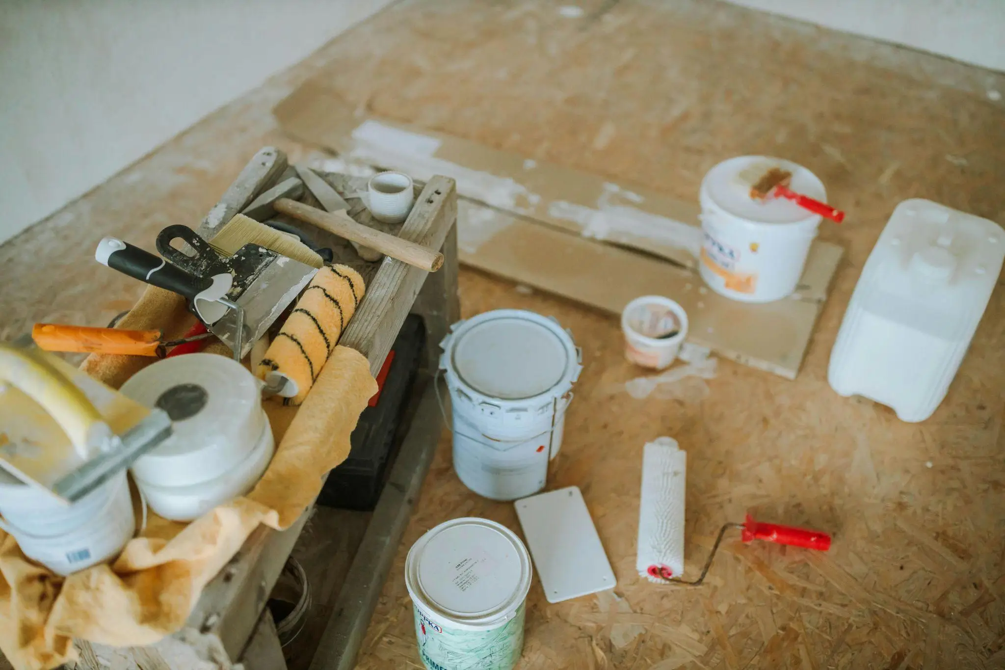 Paint supplies and tools scattered on plywood floor in a room under renovation