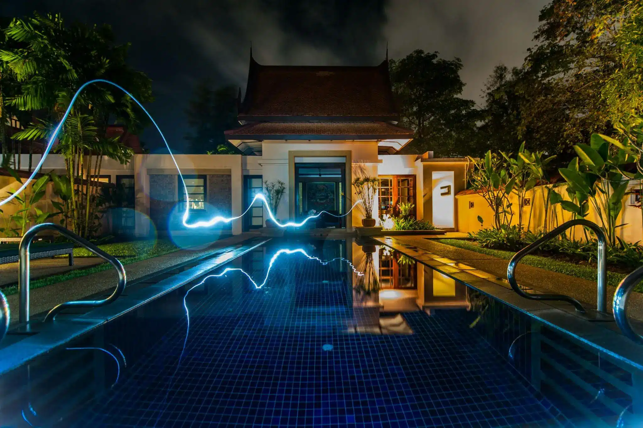 Traditional Thai house with illuminated pool and palm trees at night, light painting reflecting in water