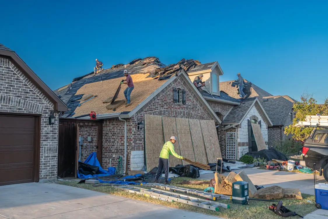 Workers replacing roof shingles on a red brick house under clear blue sky