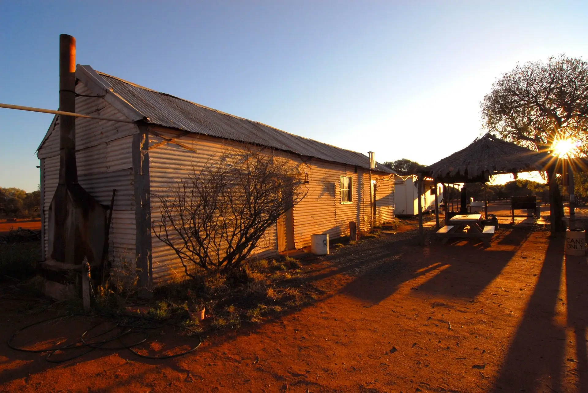Rustic corrugated metal building with thatched shelter in arid landscape at sunset