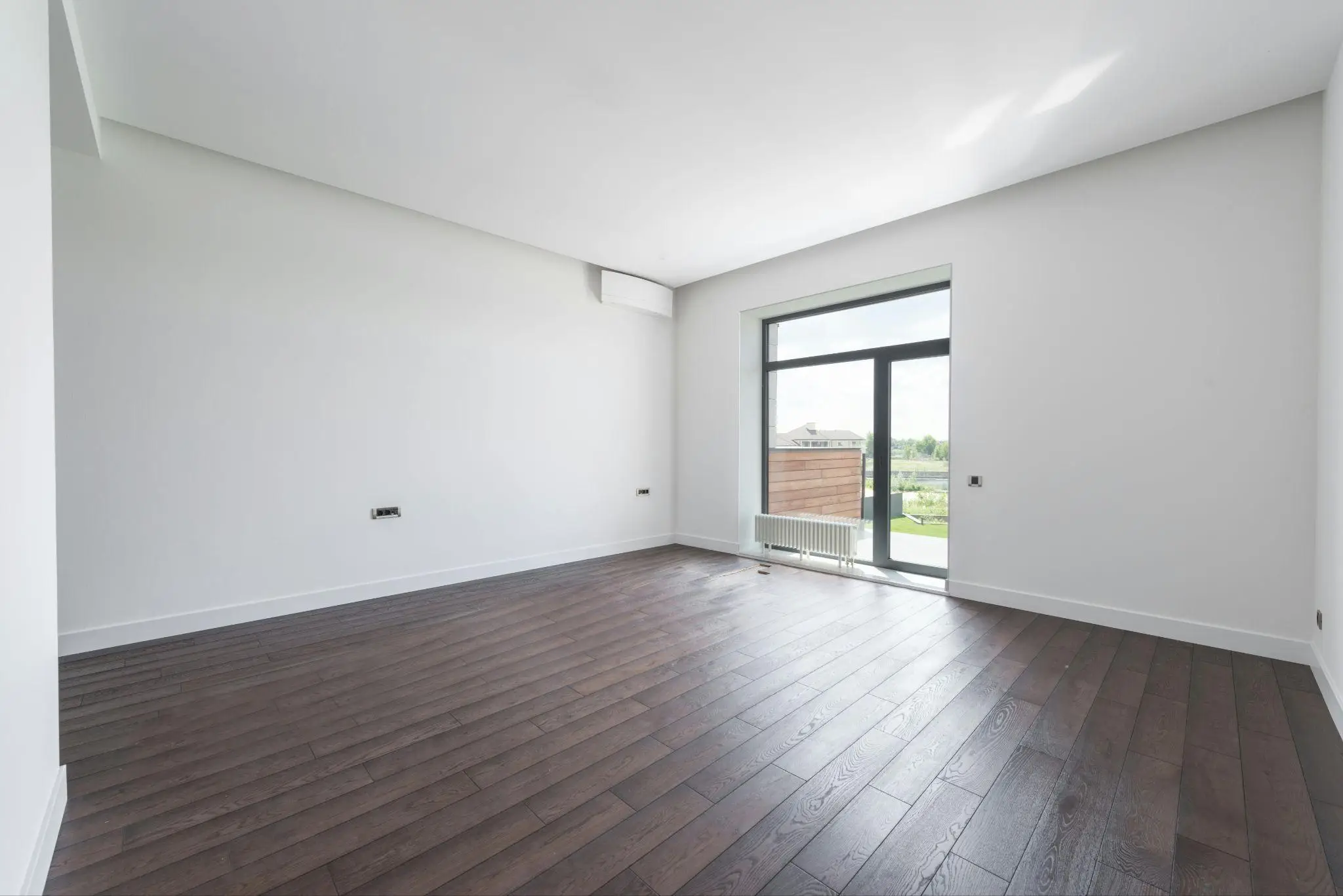 Spacious empty room with dark wooden flooring and large window overlooking a garden