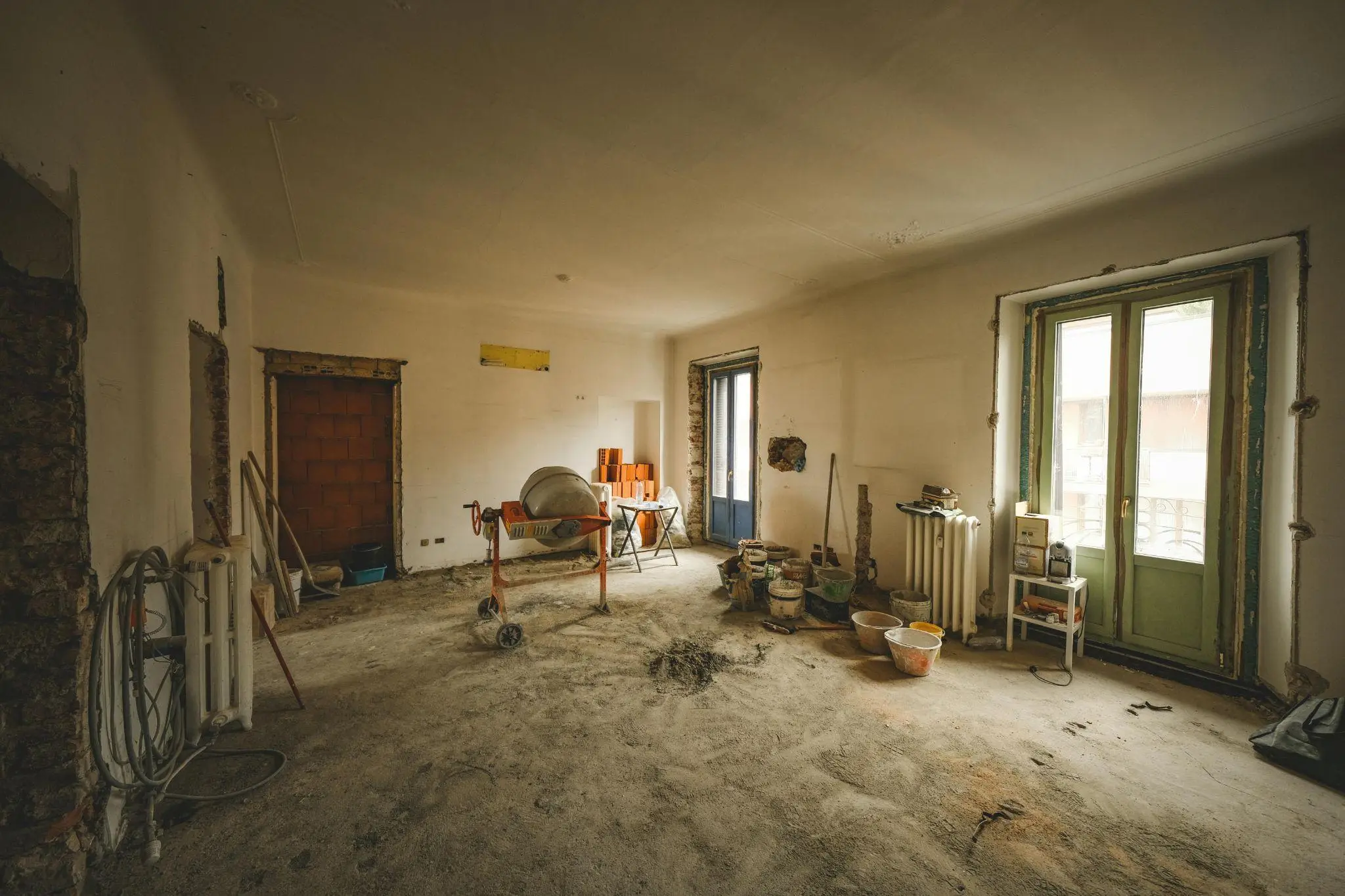 Renovation site with cement mixer, construction tools, and open balcony door in dimly lit room
