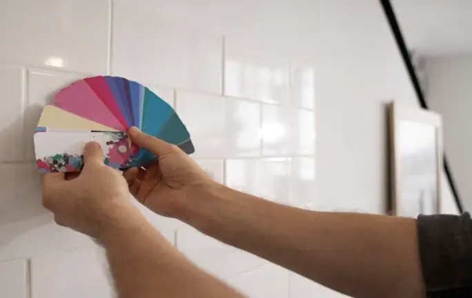 Person holding a color swatch fan against white tiled wall in well-lit room