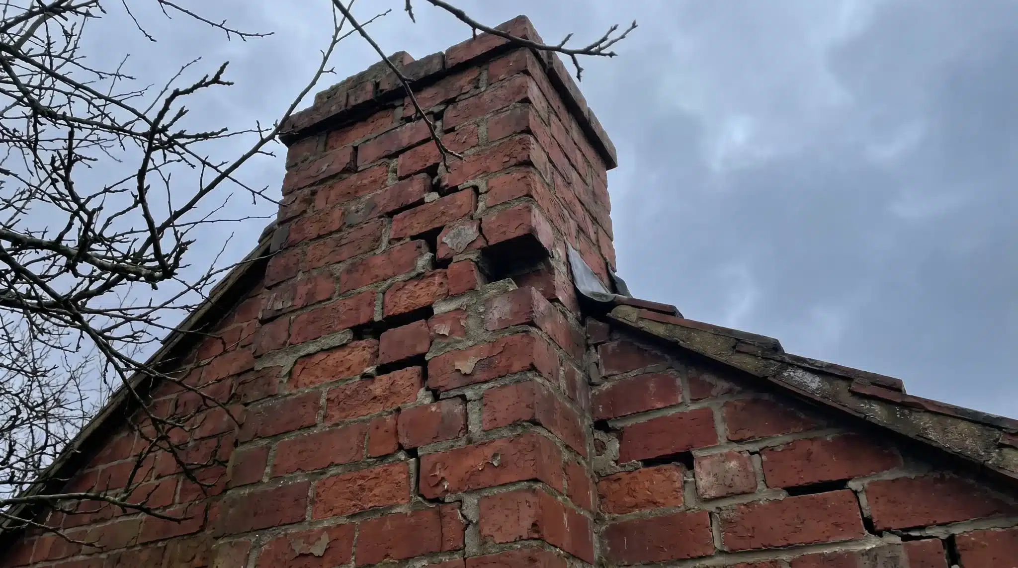 Cracked red brick chimney against cloudy sky with bare branches nearby