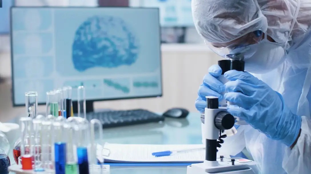 Scientist in protective gear using microscope in laboratory with test tubes and computer screen