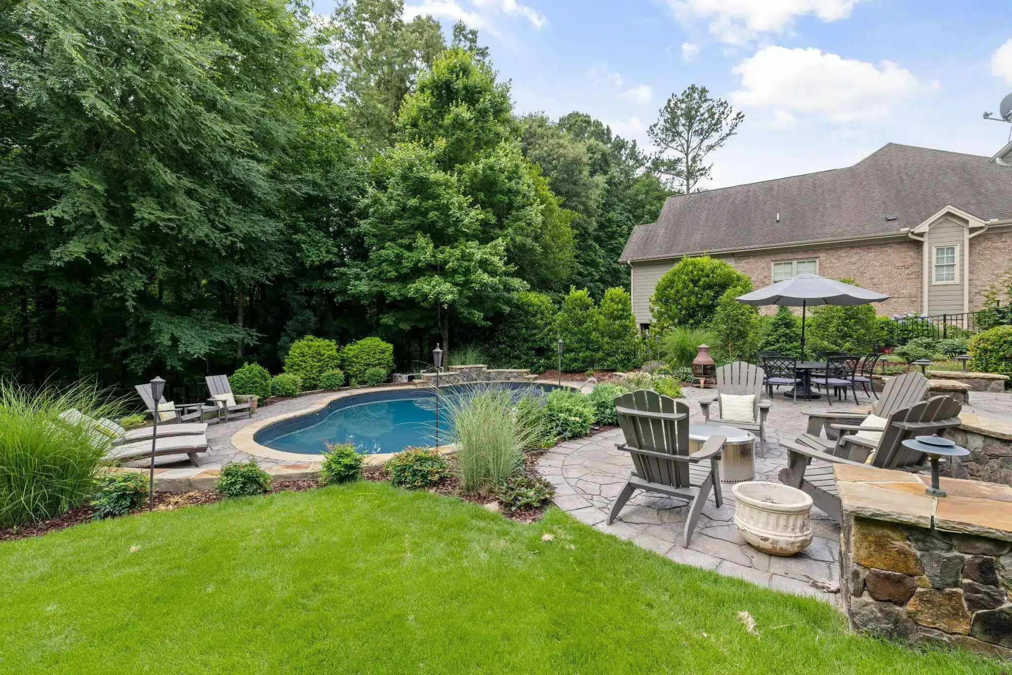 Outdoor patio with gray Adirondack chairs, a stone fire pit, and a swimming pool surrounded by trees