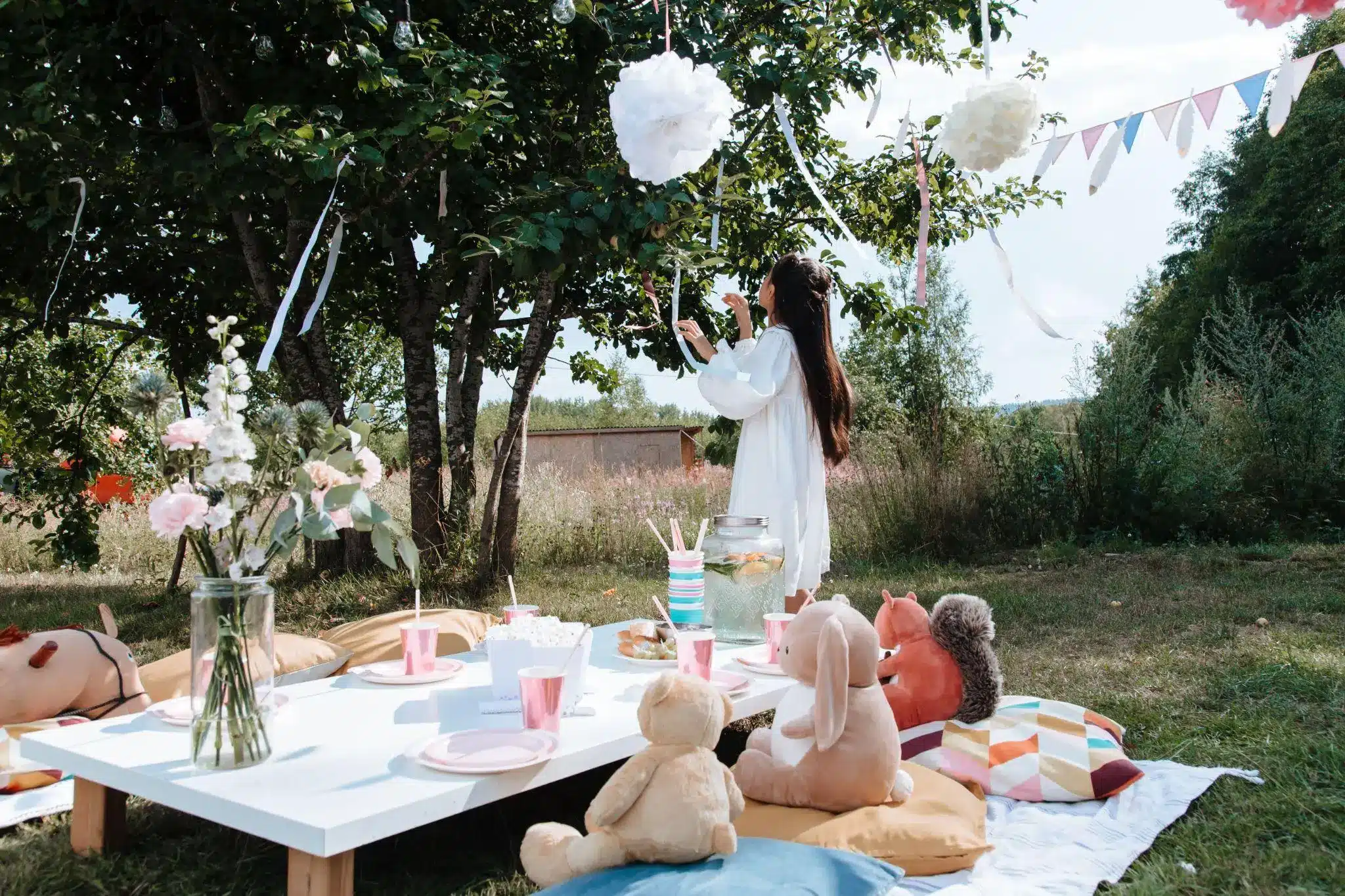 Outdoor picnic setup with stuffed animals and flowers under a tree in bright daylight