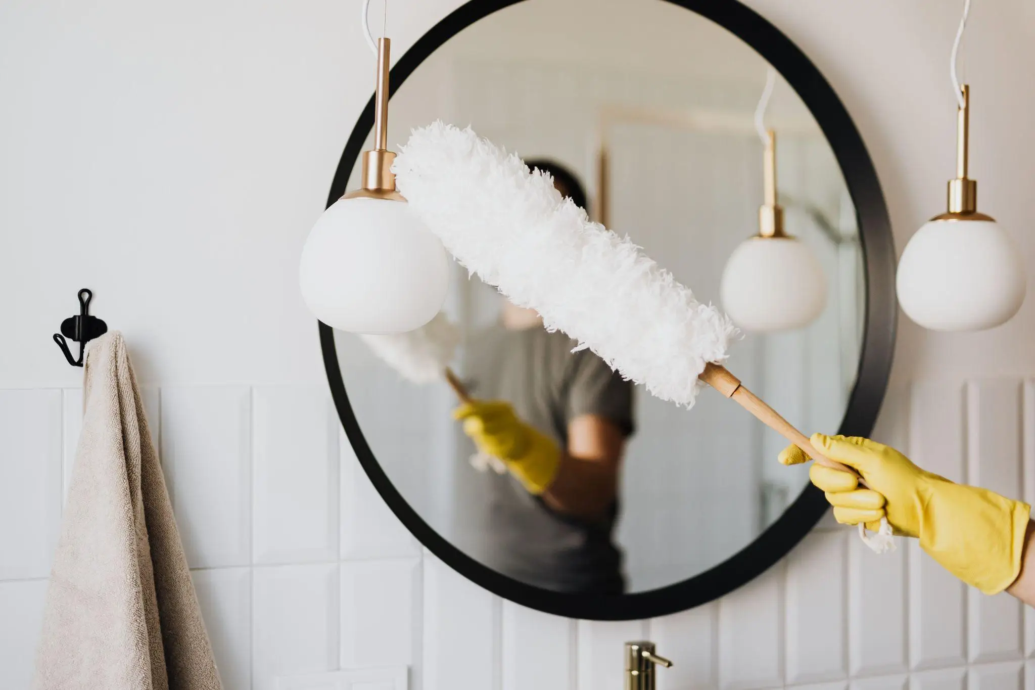 Hand in yellow glove dusting round mirror with feather duster in bathroom setting