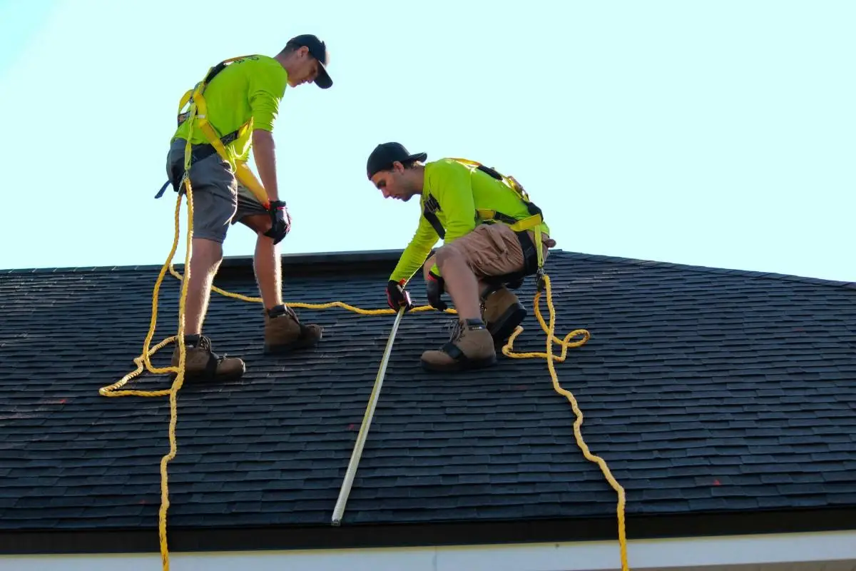 Two workers in safety gear installing shingles on a roof under clear sky