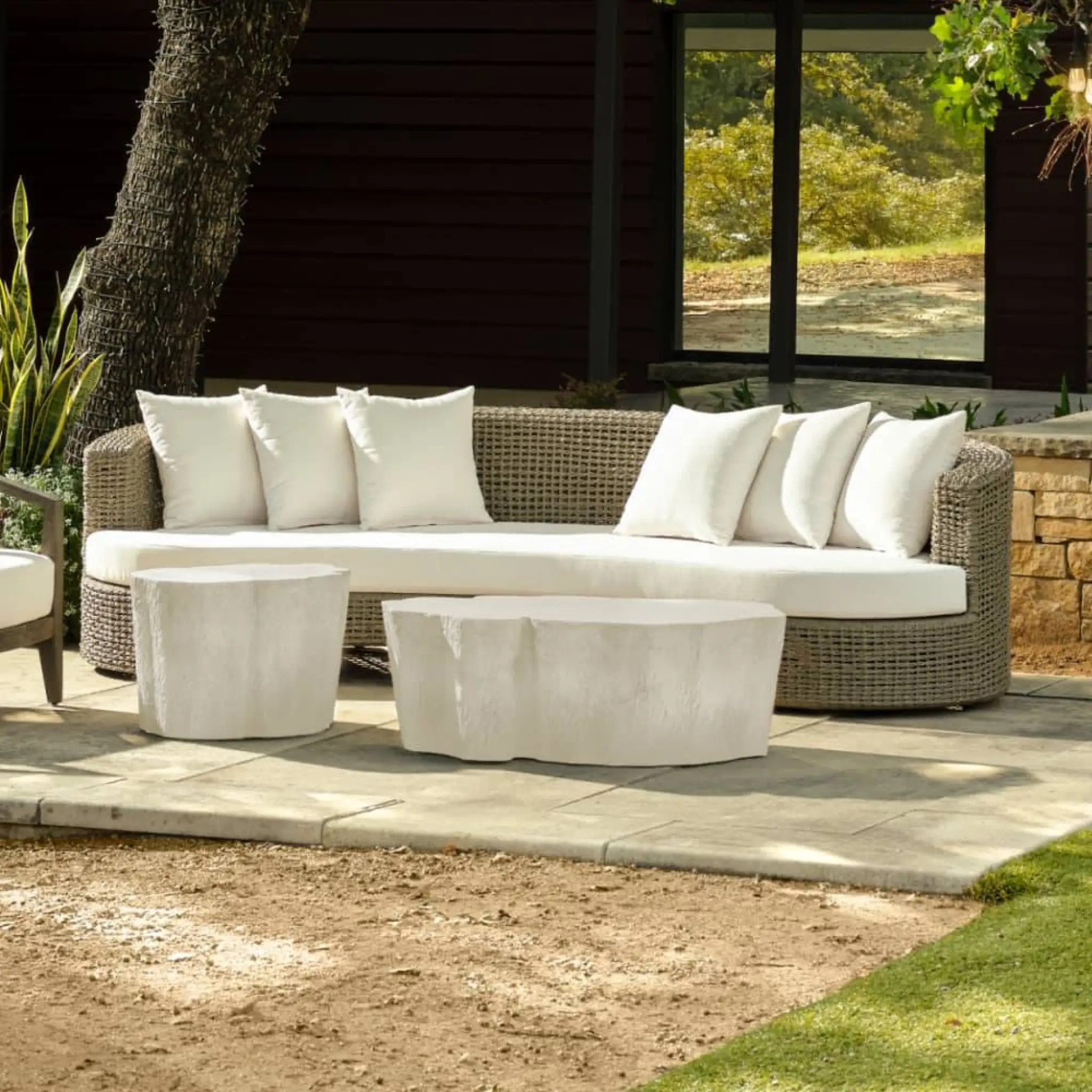 Wicker outdoor sofa with white cushions on patio near tree and decorative stools