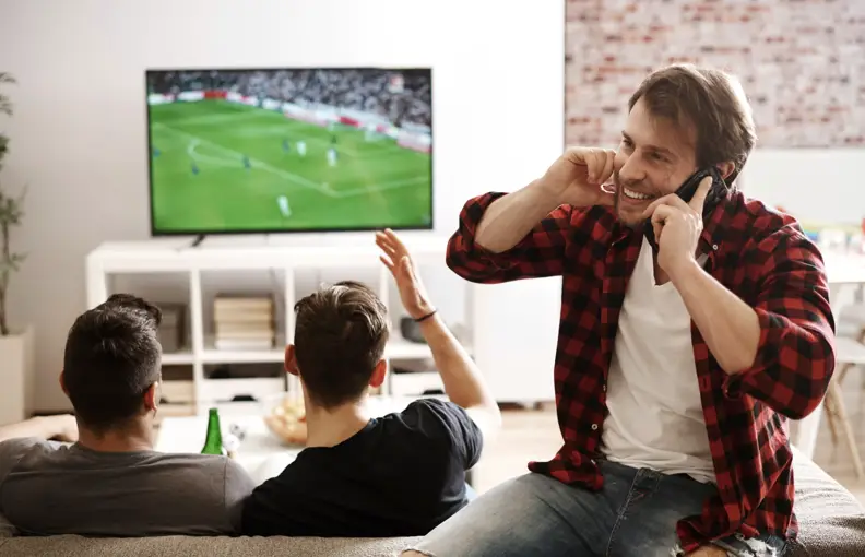 Three men watching a soccer game on TV in a living room, one on the phone