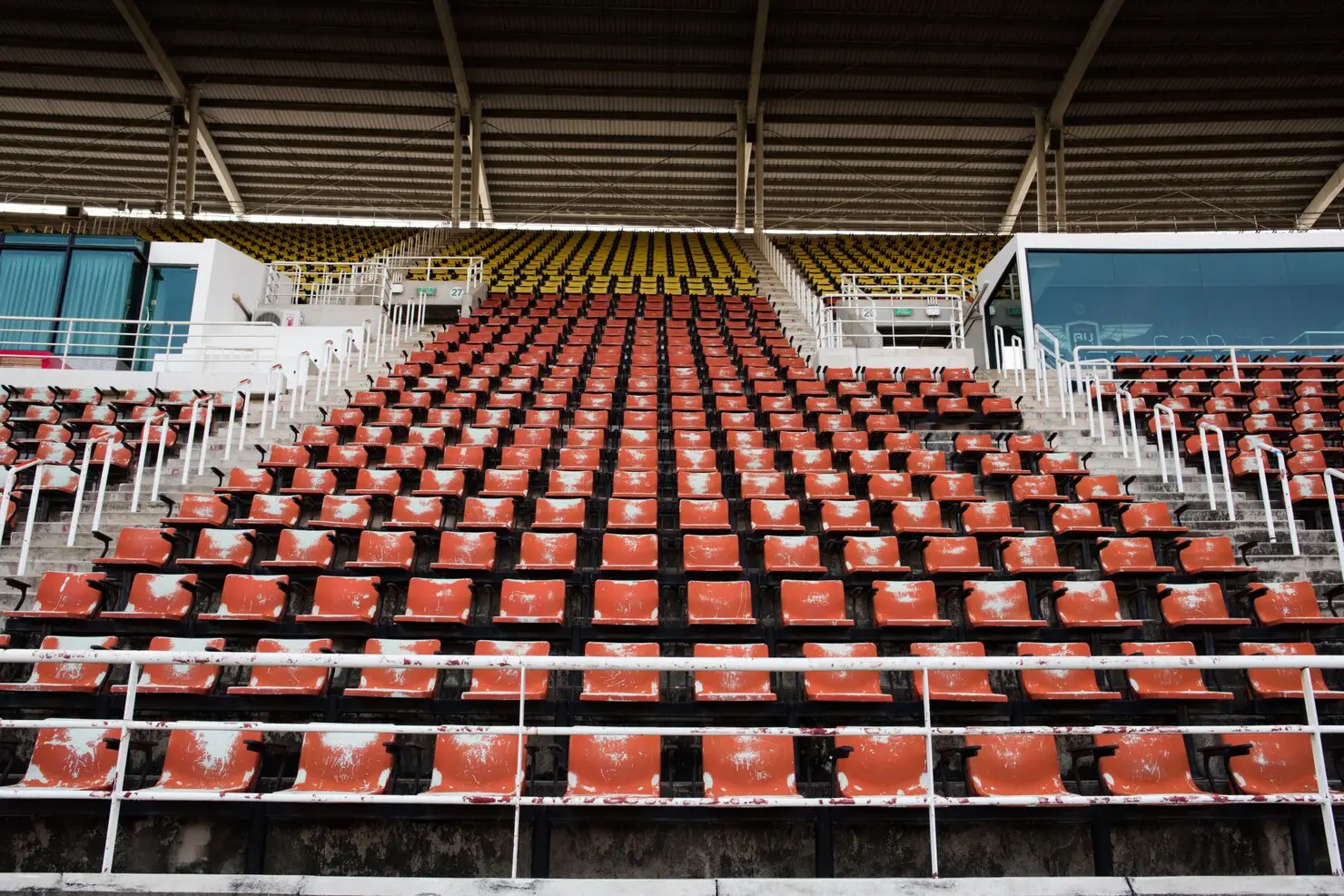 Rows of empty red and yellow stadium seats under metal roof, viewed from field level