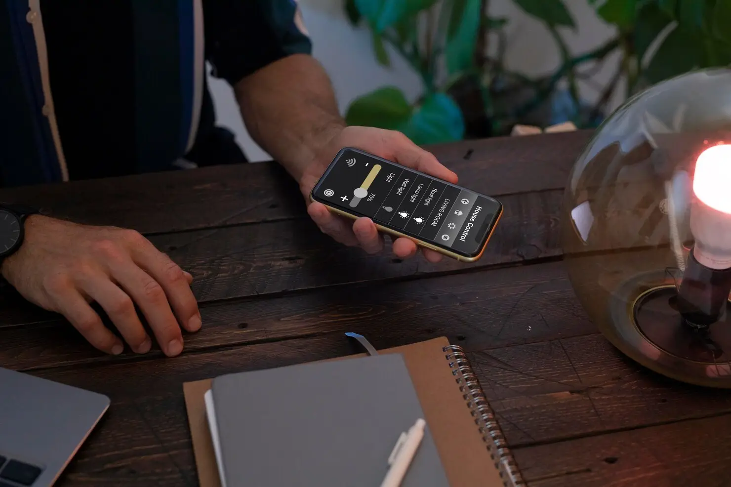 Hand holding smartphone with smart home app in dimly lit room on wooden table