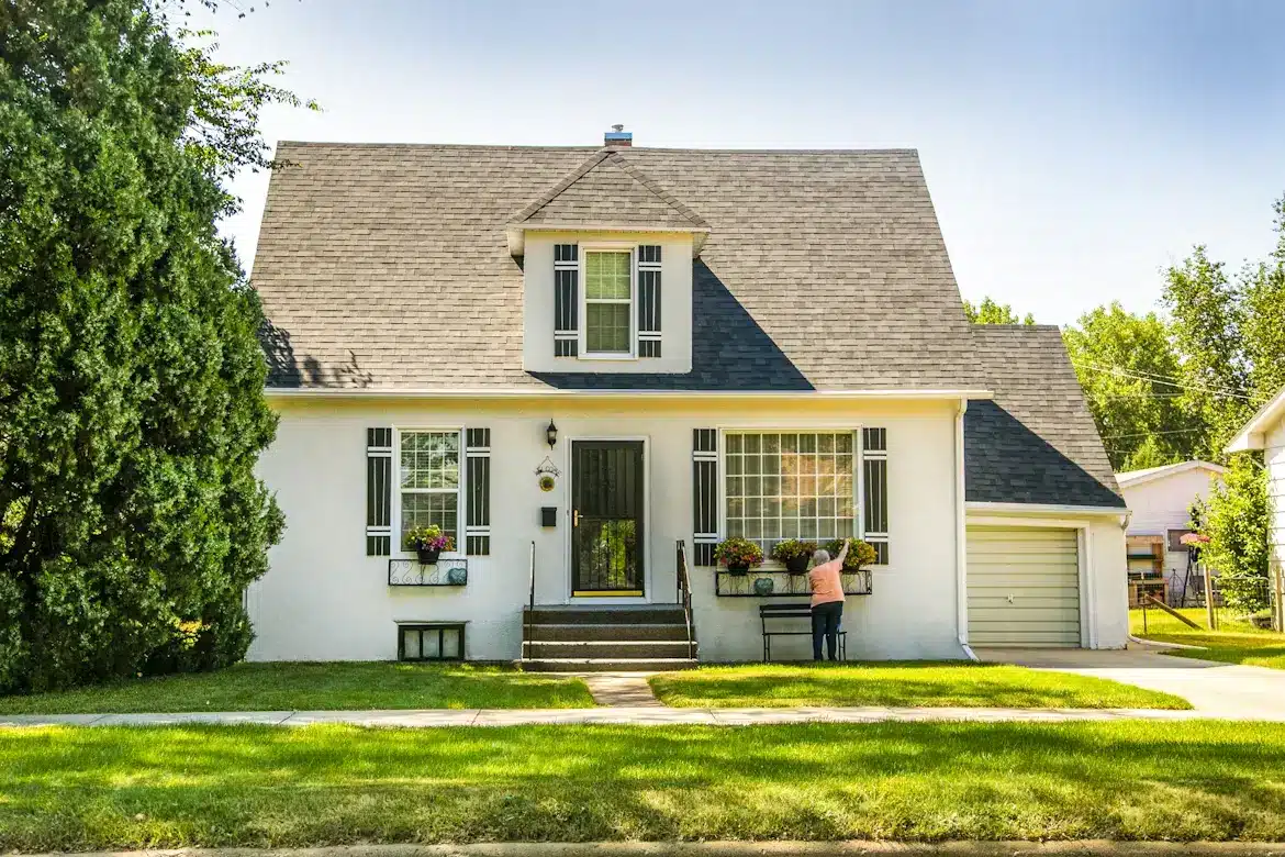 Person tending flower boxes on suburban house exterior with gable roof and white facade