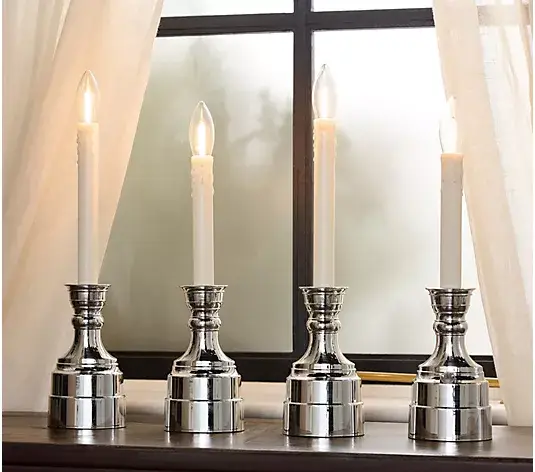 Four lit candles in shiny silver candlesticks on a wooden mantle with sheer curtains