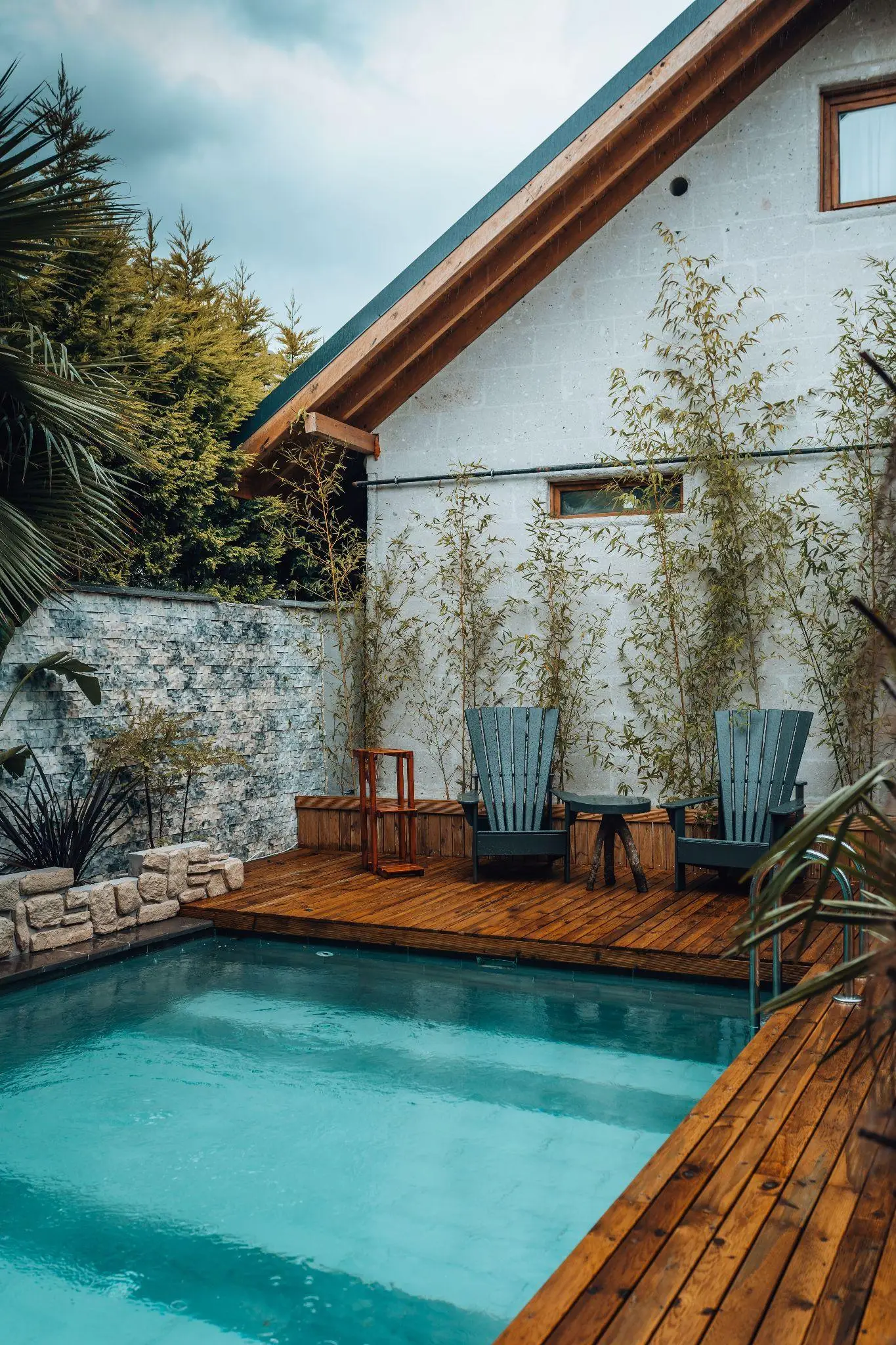 Outdoor pool area with wooden deck, two chairs, and lush greenery beside a modern house