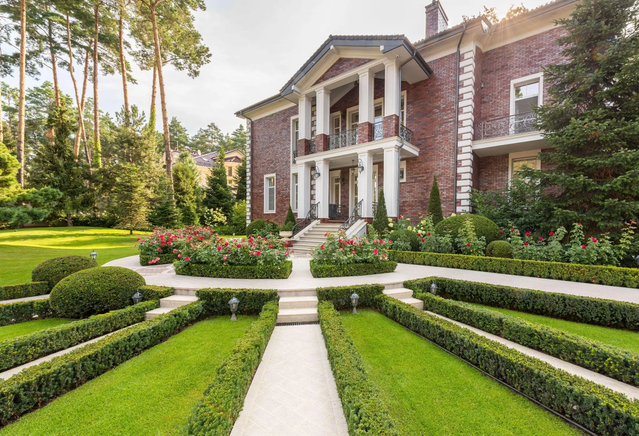 Elegant brick mansion with columned entrance surrounded by manicured garden and lush green lawn