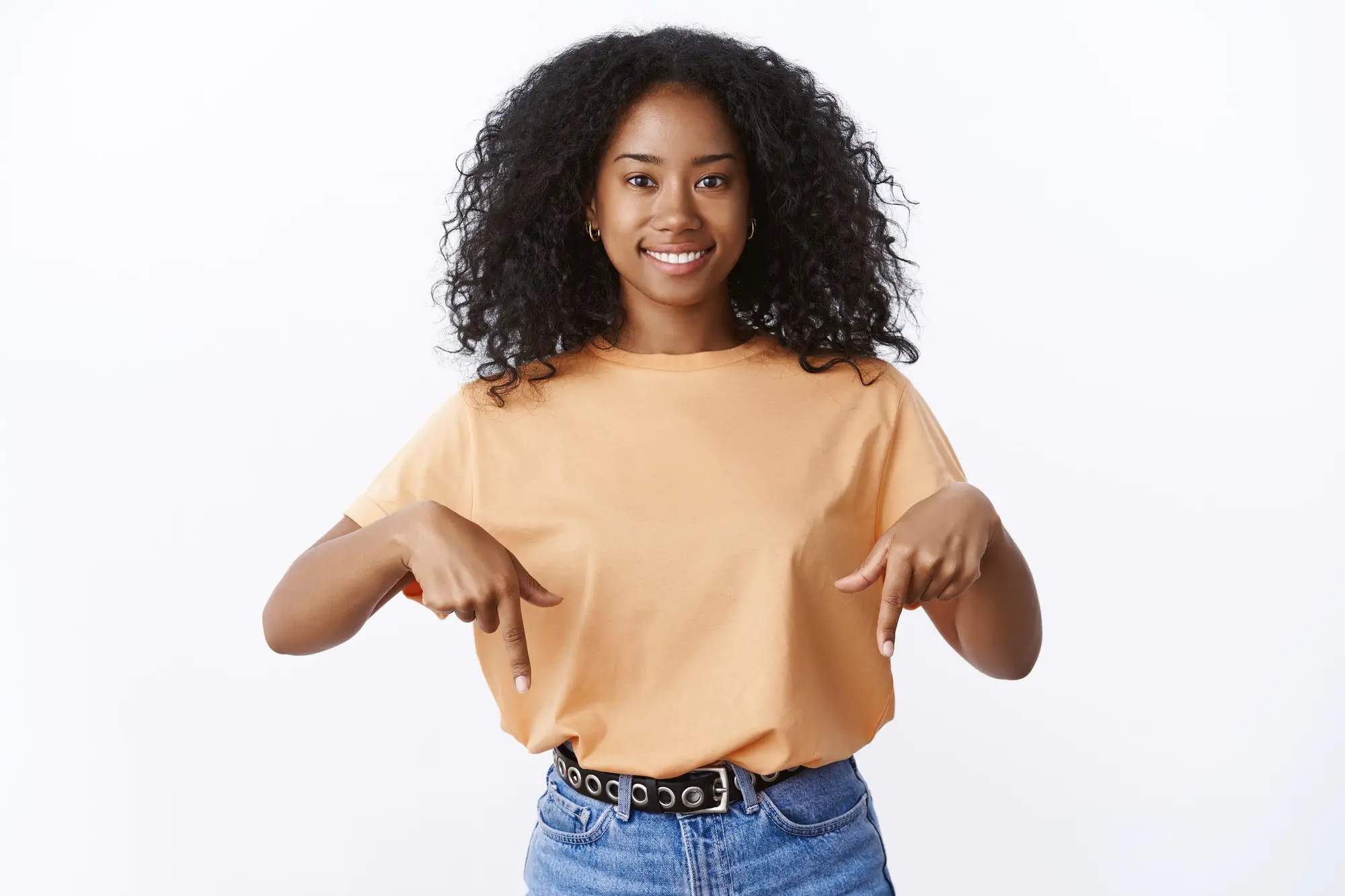 Smiling woman in a peach t-shirt pointing down with both hands against white background