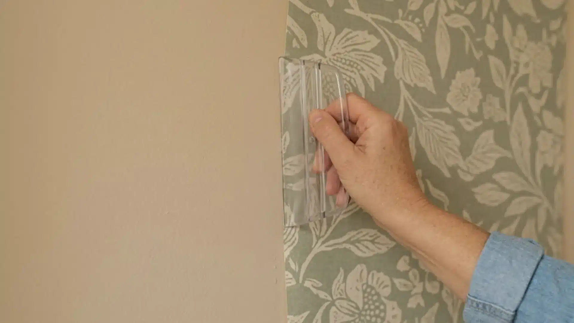 a person pressing a clear smoothing tool over sage green floral wallpaper near the wall edge