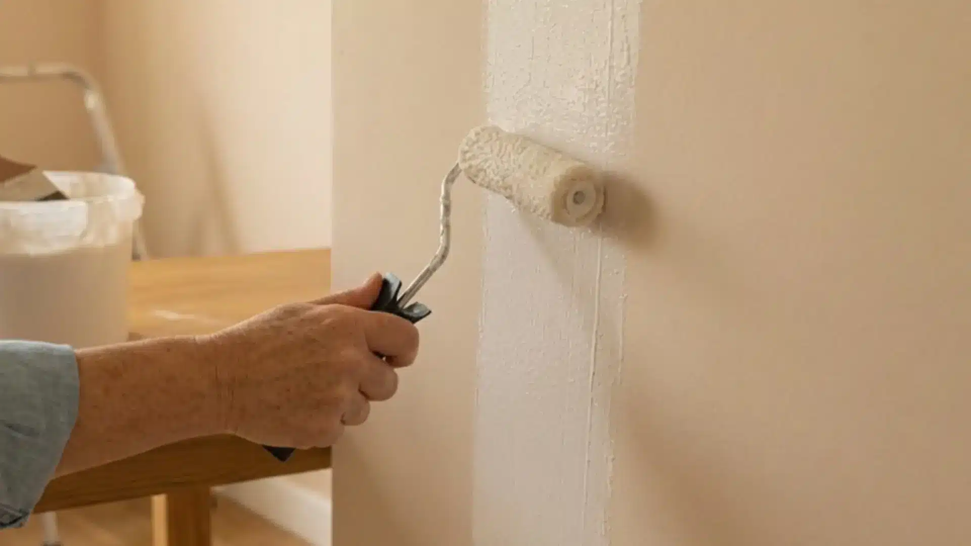 a person rolling white adhesive paste onto a beige wall using a paint roller attached to a long handle