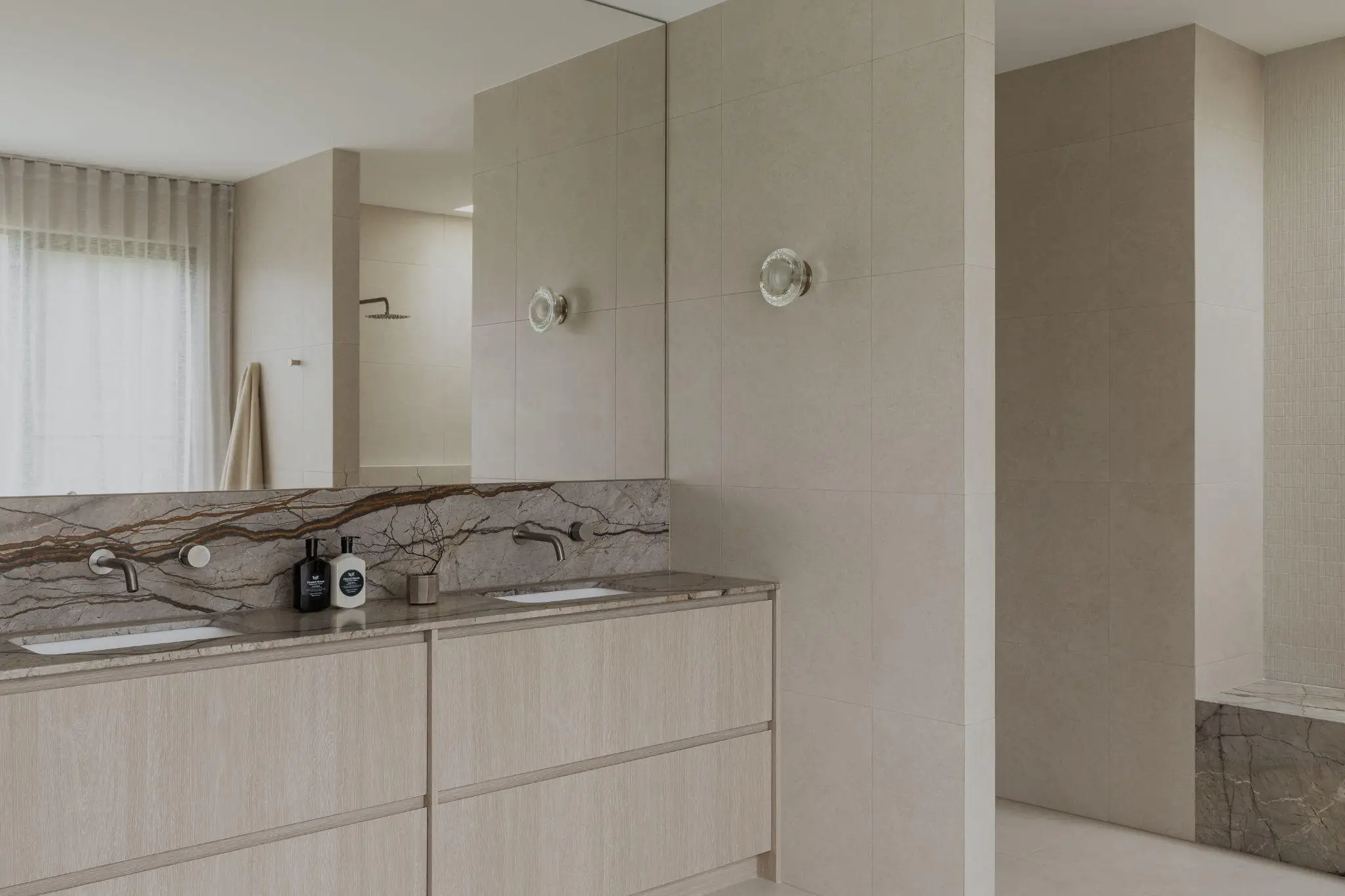 Modern bathroom with double vanity and marble countertops, beige tiles, and a glass shower enclosure