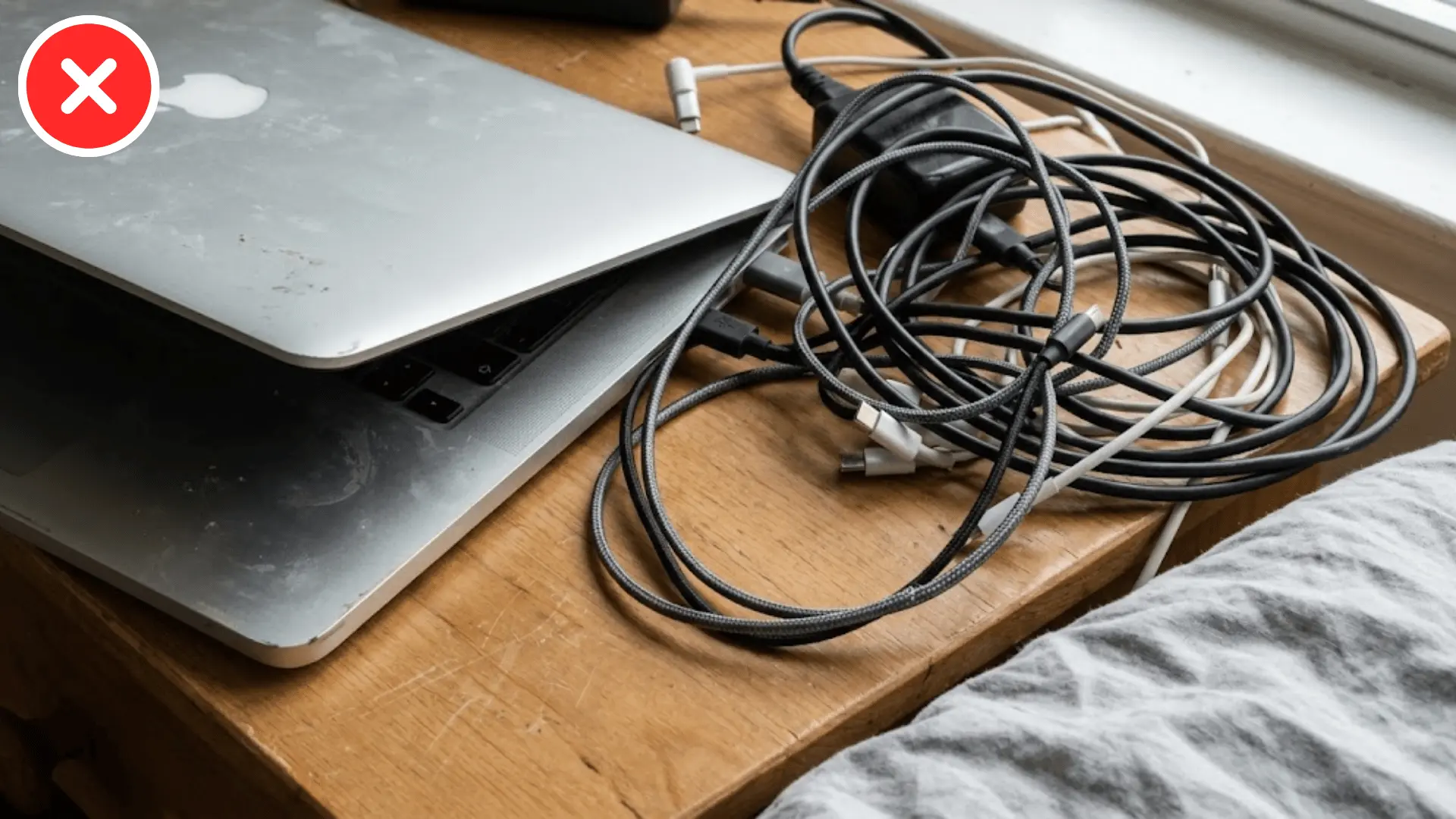 close up of a laptop and tangled cables on a desk beside the edge of a bed