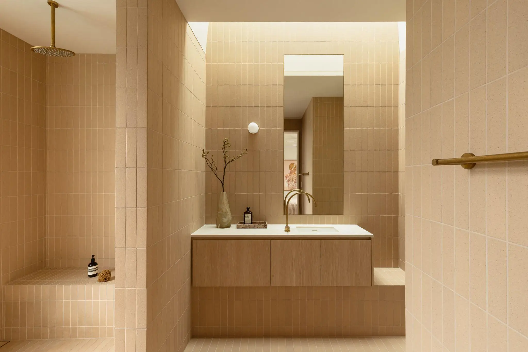 Modern beige tiled bathroom with brass fixtures and a minimalist countertop with a vase and soap dispenser