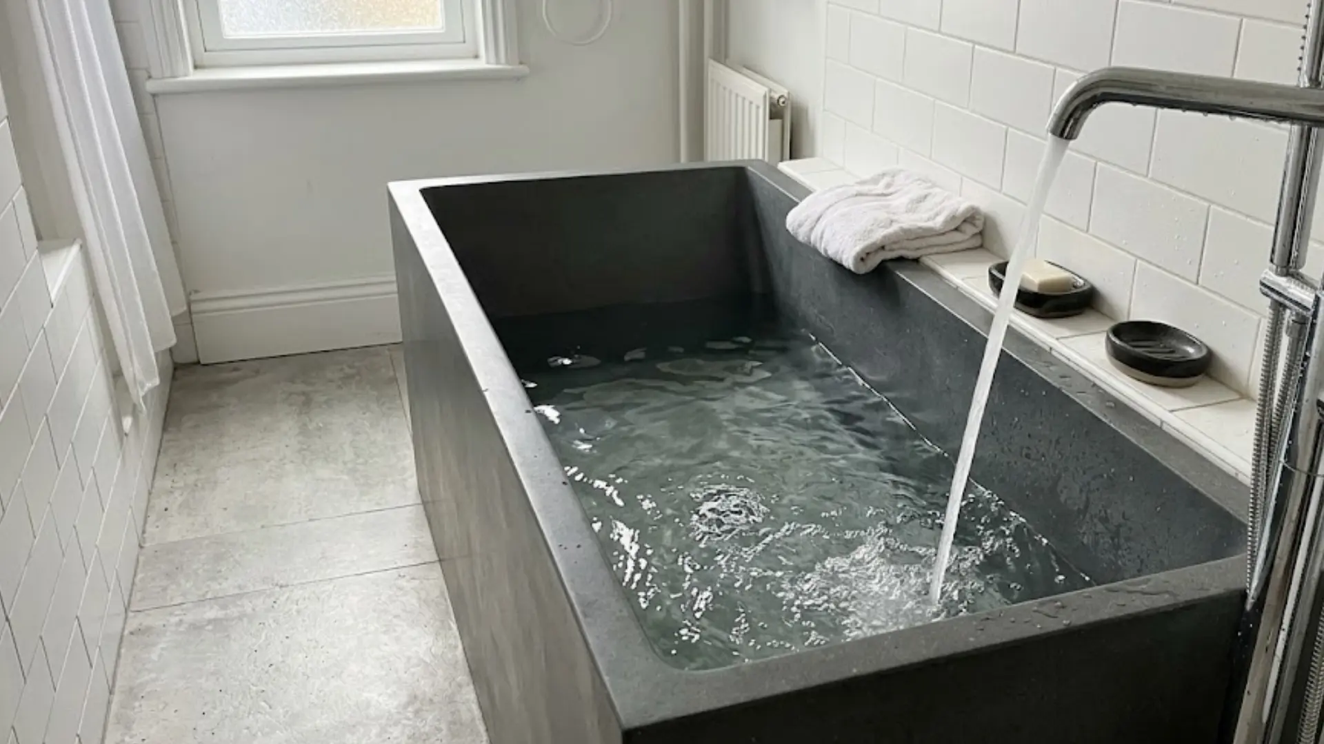 dark stone soaking tub filling with water beside folded towels and chrome faucet in tiled bathroom