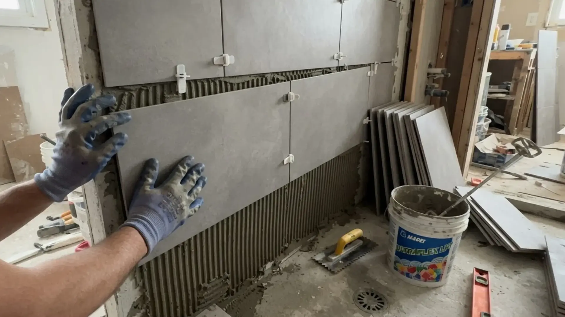 gloved hands pressing a large gray tile onto a shower wall coated with thinset mortar