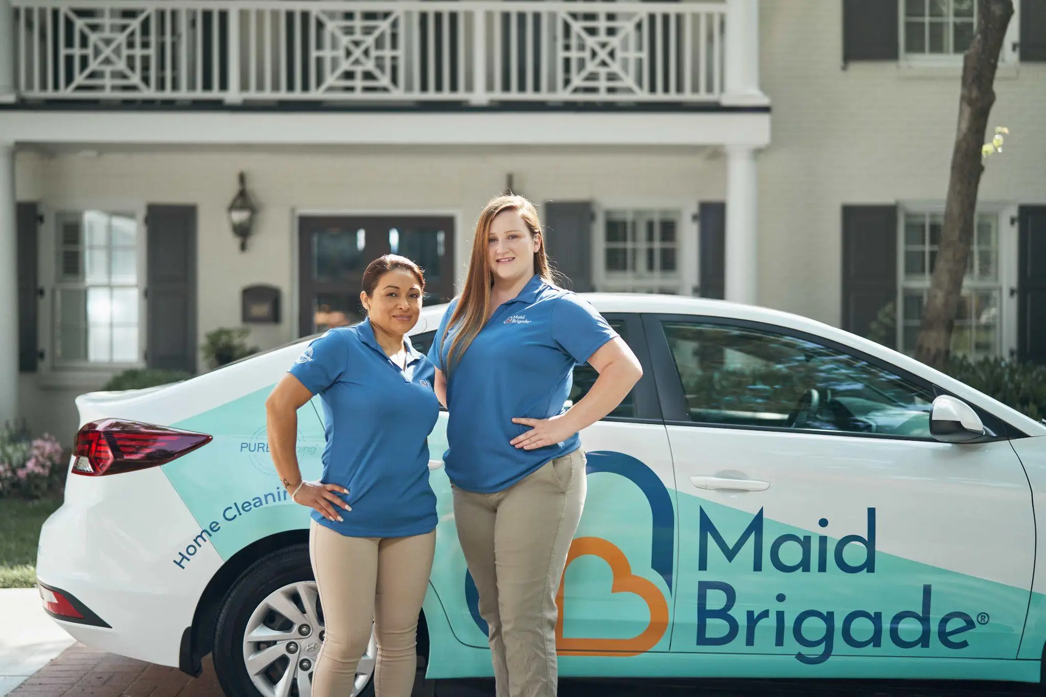 Two women in blue uniforms standing beside Maid Brigade car in residential driveway
